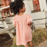 A young child wearing a peach pink cotton gauze dress with white lace trim, standing outdoors in front of a white wooden house.