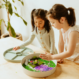 Two young girls playing with a small garden setup on a table.