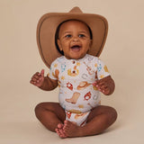A smiling baby sitting cross-legged wearing a tan cowboy hat and a white short-sleeve bodysuit with a western-themed print of boots, horseshoes, stars, and a guitar, photographed against a solid beige studio background.