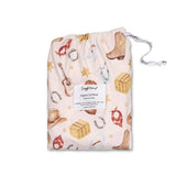 A drawstring storage bag containing an organic cotton sheet with a western-themed print, including boots, hats, horseshoes, and stars, on a white background.