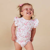 A baby girl wearing a white short-sleeve bodysuit with a pink flamingo and floral print, standing against a solid beige background.