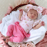 A sleeping newborn baby wearing a light pink floral short-sleeve romper and bright pink ribbed pants, styled in a wicker basket with white and pink blankets.
