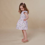 A young girl with a pink hair bow stands barefoot against a solid beige studio background, wearing a sleeveless white dress with a colorful floral and butterfly print.