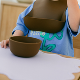 Child wearing a bib and blue shirt with green text, sitting at a table with a brown bowl.