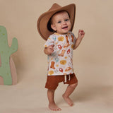 A baby wearing a white short-sleeve t-shirt with a western-themed print (boot prints, cowboy hats, and stars), matching brown shorts, and a brown cowboy hat, standing barefoot against a beige background.