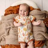 A baby lying on a cozy knit blanket and rust-colored fabric, wearing a short-sleeve footless romper with a warm-toned space-themed print.