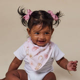 A smiling baby wearing a white ribbed short-sleeve bodysuit and a light pink patterned bib, with dark hair styled in two pigtails using pink bows, posed against a solid beige studio background.