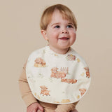 A smiling baby wearing a cream-colored bib with a cute print of teddy bears, puppies, and a small duck, paired with a beige long-sleeve top, set against a solid beige background.