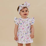 A toddler girl wearing a white short-sleeve dress with a colorful floral print and a matching bow headband, standing against a solid beige background.