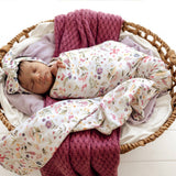 A sleeping newborn baby swaddled in a white floral blanket with a matching floral bonnet, resting in a woven basket with a dusty pink knit blanket.
