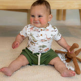 Baby sitting on a carpeted floor wearing a dinosaur-themed shirt and green shorts, holding a brown toy.