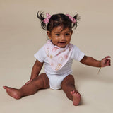 A baby girl sitting on a beige studio background, wearing a white short-sleeve bodysuit, a light pink patterned bib, and pink hair bows.