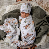 A sleeping newborn baby swaddled in a white wrap blanket with a brown and green western print, wearing a matching hat. The baby is nestled in a wicker basket lined with a dark olive green blanket and a cream knit blanket.