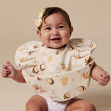 A smiling baby wearing a cream bib with a bee and flower pattern, set against a solid beige background.
