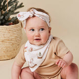 A baby wearing a cream-colored fabric headband and matching bib with a rustic holiday print, paired with a beige short-sleeve romper. The baby is posed next to a woven basket containing evergreen branches against a solid beige background.