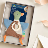 A duck-shaped fabric teething toy with a ring and colorful feather accents, presented inside a white gift box with its retail card visible behind it. The toy has a green head, blue wings, brown body, and orange feet.