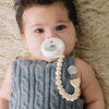 Baby wearing a gray knit outfit with a white pacifier and beige teething ring on a beige textured surface