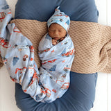 Newborn baby swaddled in a car seat-themed blanket and hat, lying on a textured surface.