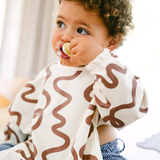 Child wearing a white bib with brown wavy patterns, holding a pacifier.