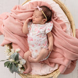 A sleeping newborn baby wearing a white short-sleeve bodysuit with a pink flamingo and floral print, resting in a woven basket with a pink knit blanket and a pink bow headband.