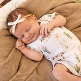 A sleeping newborn baby wearing a white short-sleeve bodysuit with a desert cactus and rabbit print, a white bow headband, and lying on a textured brown blanket.