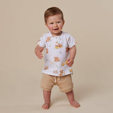 A toddler standing barefoot against a plain beige studio background, wearing a white short-sleeve top with a teddy bear print and beige shorts.