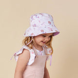 A young girl wearing a light pink and white mermaid-themed bucket hat and a pale pink overall dress, photographed against a solid beige background.