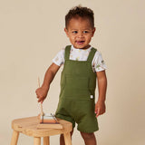 A toddler stands next to a small wooden stool wearing olive green overall shorts over a white t-shirt with a tropical print. The child is holding a wooden toy mallet on a wooden xylophone.