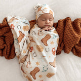 A sleeping newborn baby swaddled in a cream-colored western print swaddle blanket and matching beanie hat, lying on a white bed with a textured rust-brown knit blanket.