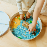 Children playing with a sensory bin filled with colorful sand and small toys on a wooden floor.