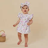 A toddler girl wearing a white floral print dress with ruffled sleeves and a matching bow headband, standing barefoot against a solid beige background.