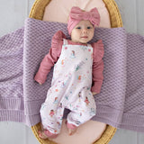 Baby in pink outfit with floral overalls and headband, lying on a textured blanket.