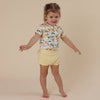 Child wearing a colorful shirt with fruit pattern and yellow shorts on a beige background