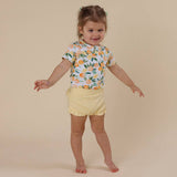 Child wearing a colorful shirt with fruit pattern and yellow shorts on a beige background