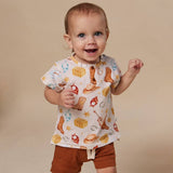 A smiling toddler stands against a plain beige studio background wearing a short-sleeve white top with a colorful western print and brown shorts.