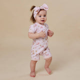 A baby girl wearing a light pink short-sleeve romper with a bunny print and a matching pink bow headband, standing barefoot against a solid beige background.