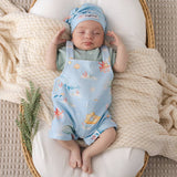 A sleeping newborn baby wearing a light blue short-sleeve romper and matching beanie with ocean-themed prints, lying on a white cushion inside a wicker basket with a cream knit blanket.