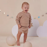 Baby in a beige outfit standing in front of a cloud and star decorated wall.
