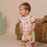 A toddler stands in a styled studio setting wearing a white short-sleeve romper with a western-themed print including boots, horseshoes, stars, and a horseshoe motif. The romper has brown trim at the sleeve cuffs and leg openings.