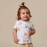 A toddler girl with a small hair bun wearing a white short-sleeve Henley t-shirt with a brown bear and yellow flower print, paired with beige shorts, against a solid beige background.