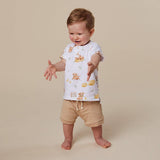 A toddler standing barefoot against a plain beige studio background, wearing a white short-sleeve T-shirt with a small teddy bear and fruit print, paired with beige shorts.