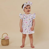 A baby girl wearing a white short-sleeve dress and matching bow headband with a colorful floral print, standing against a solid beige background next to a small wicker basket with a pink rose.