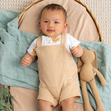 A baby lying on a quilted cream rug, wearing beige overall-style romper overalls with a white t-shirt underneath and a small beige plush bunny toy beside them.