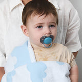 Baby with a pacifier and blue blanket, held by an adult against a gray background