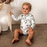 Baby sitting on a sandy surface wearing a floral long-sleeve onesie.