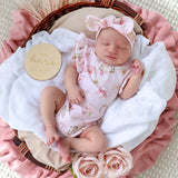 A sleeping newborn baby wearing a light pink floral romper and matching bow headband, lying in a wicker basket with white muslin cloth, pink ruffled blankets, a wooden sign reading 'I'm here', and pink roses.