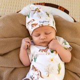 A sleeping newborn baby wearing a matching white swaddle blanket and knit hat set with a desert and horse print, resting on a textured brown blanket.