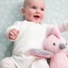 Baby in a white outfit holding a pink plush toy against a light green background with floral patterns.