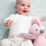Baby in a white outfit holding a pink plush toy against a light green background with floral patterns.