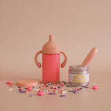 Pink sippy cup, jar with spoon, and colorful candies on a beige background